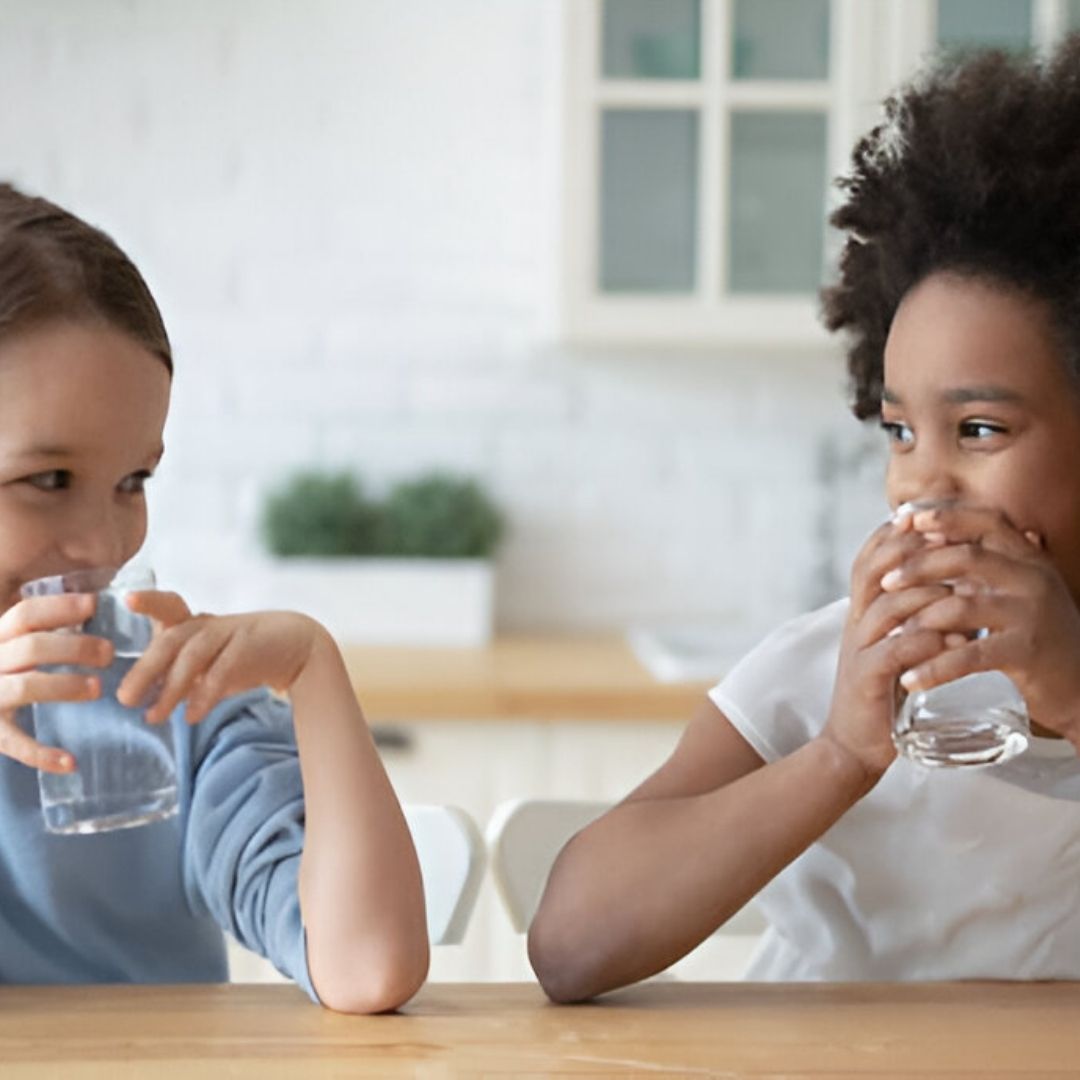 Two young girls drinking water in kitchen looking at each other